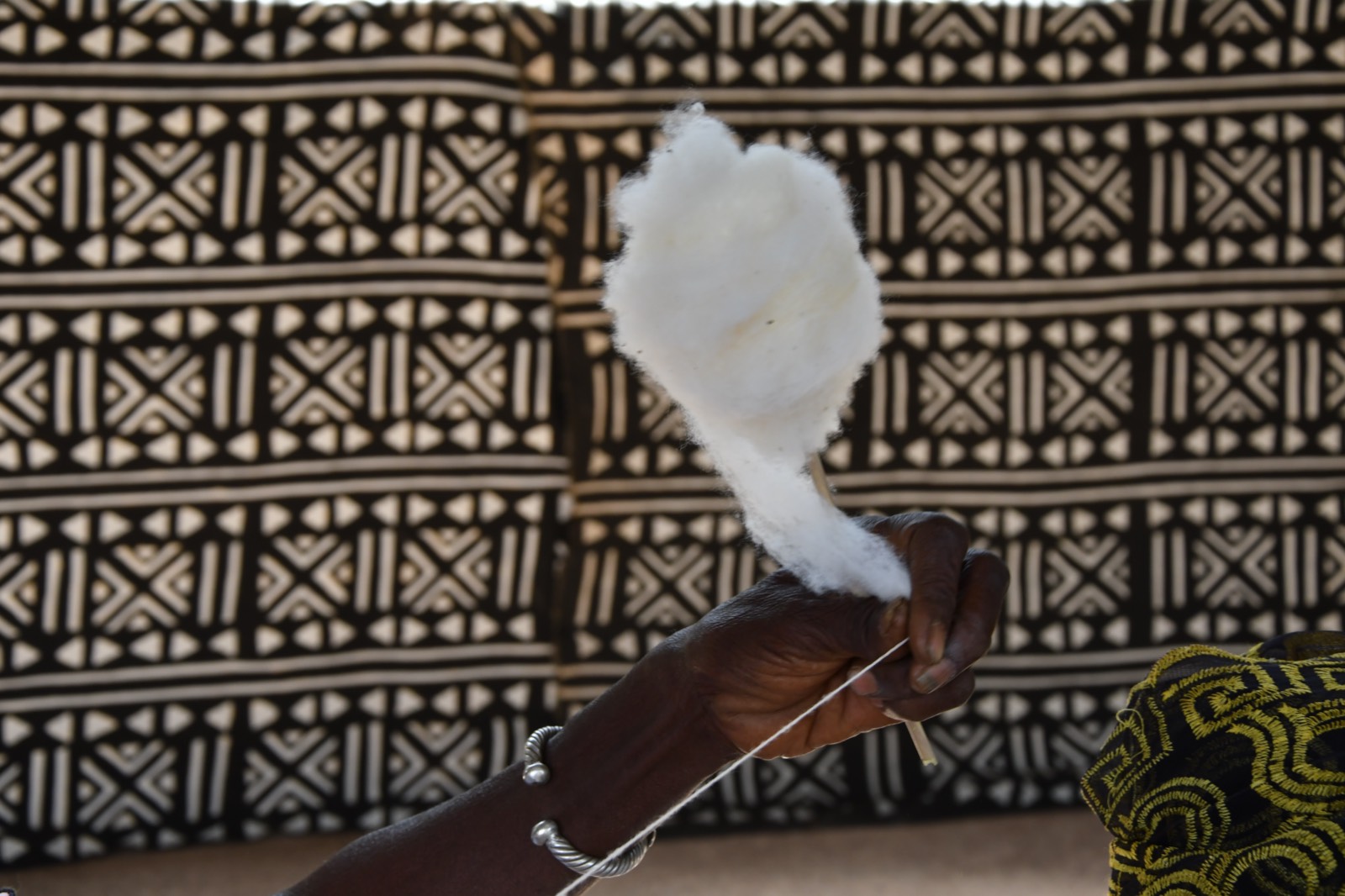 Hands spinning raw cotton into thread with a spindle, against a black-and-white bogolan mudcloth, Burkina Faso